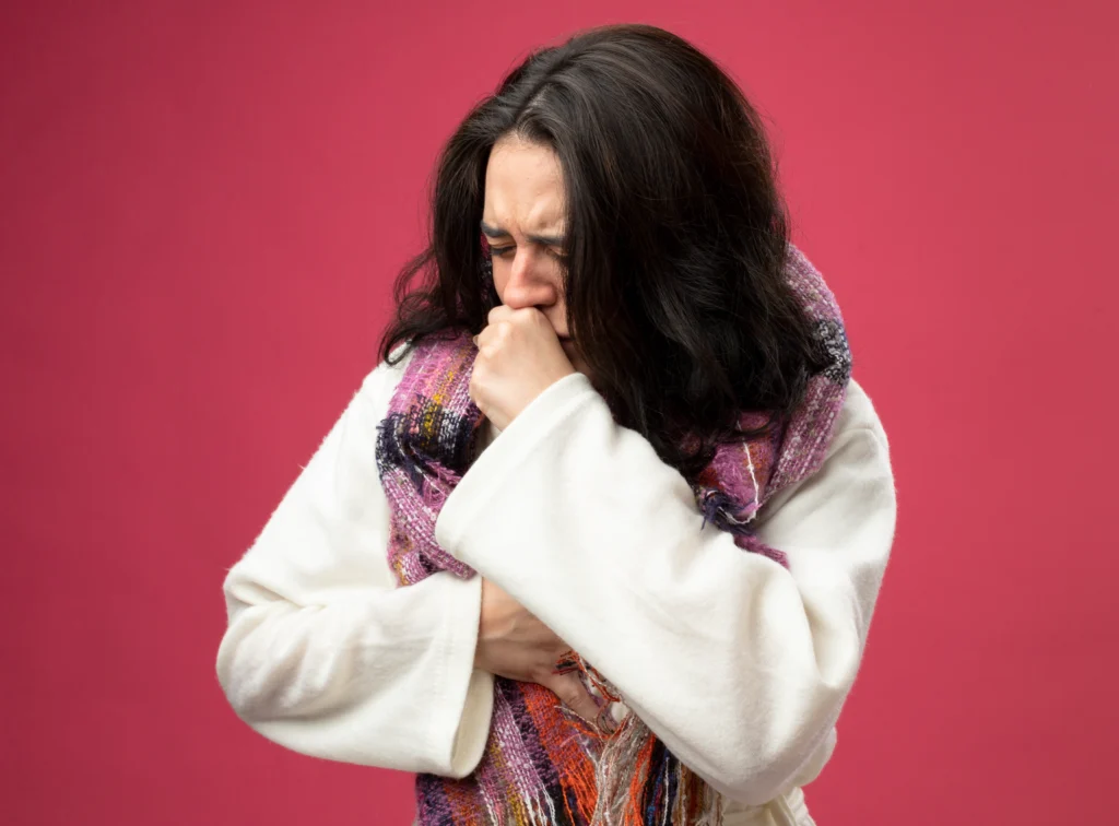 A woman wrapped in a scarf coughing into her fist against a red background.