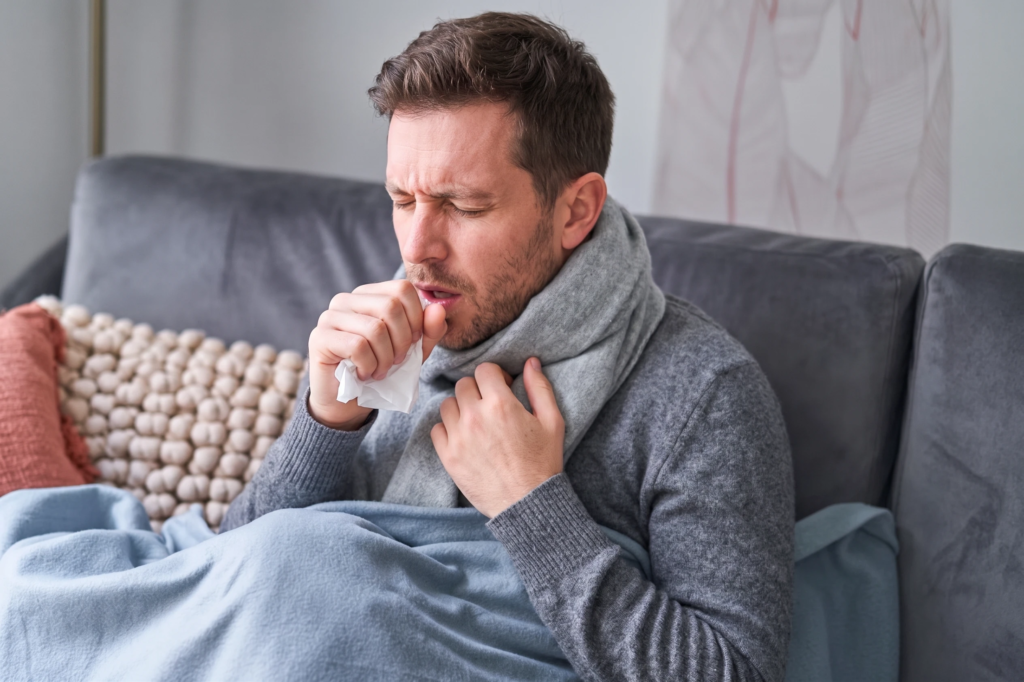 A sick man wrapped in a blanket on a couch, coughing into a tissue.