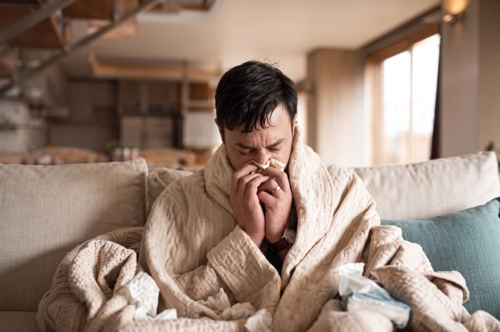 A sick man wrapped in a blanket on a couch, blowing his nose into a tissue.