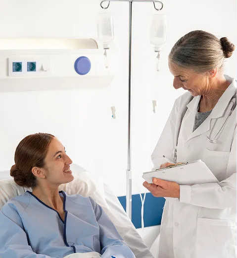 A smiling patient in a hospital bed talks with a doctor holding a clipboard.