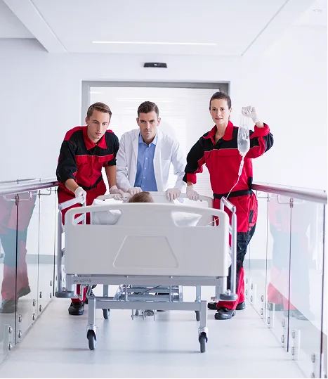 Two paramedics in red-and-black uniforms and a doctor in a white coat urgently push a hospital bed through a bright corridor.