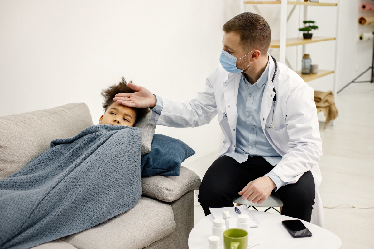 A masked doctor checking a sick child's temperature as she lies under a blanket on a couch.