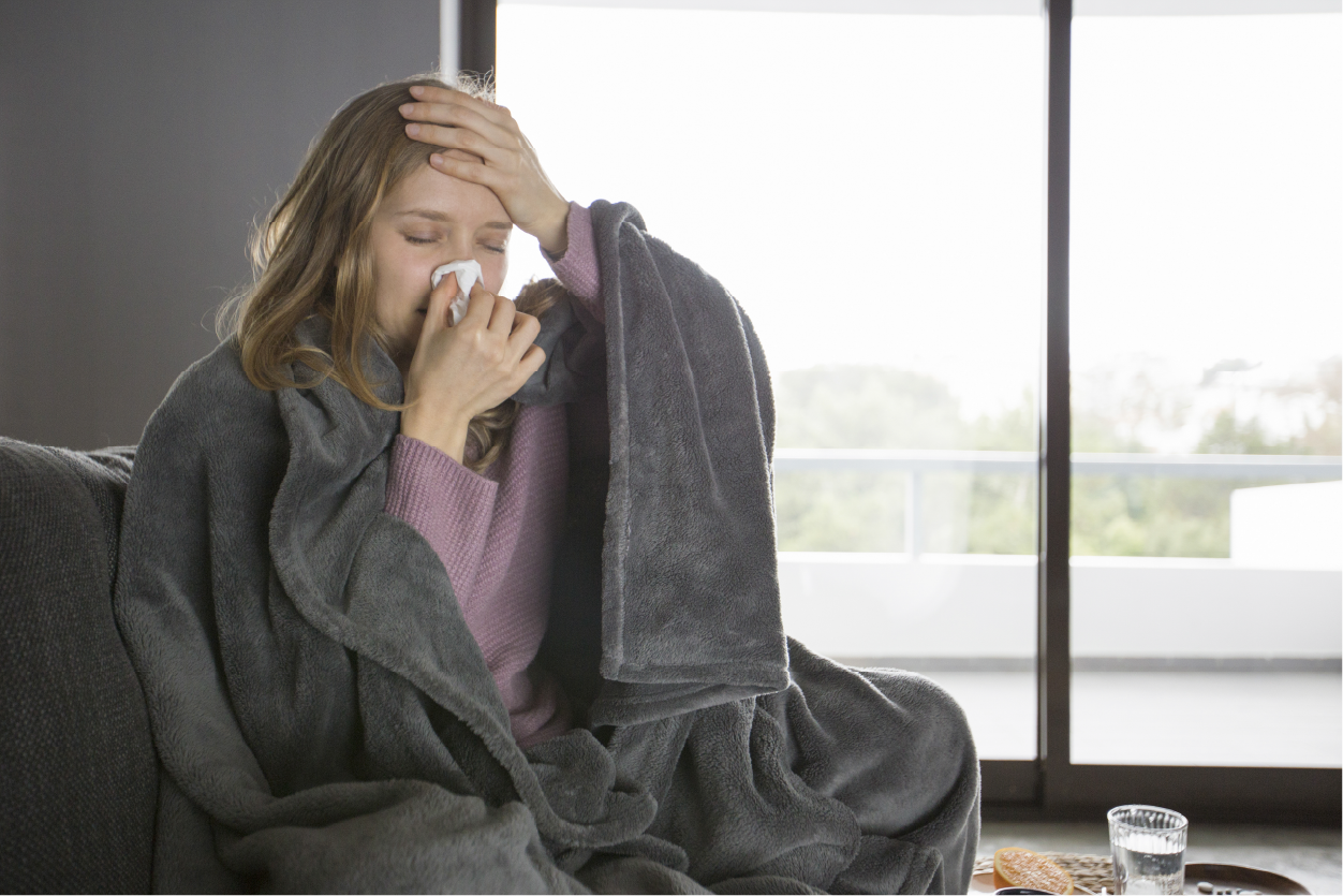 A sick woman wrapped in a blanket blowing her nose with a tissue while holding her forehead.